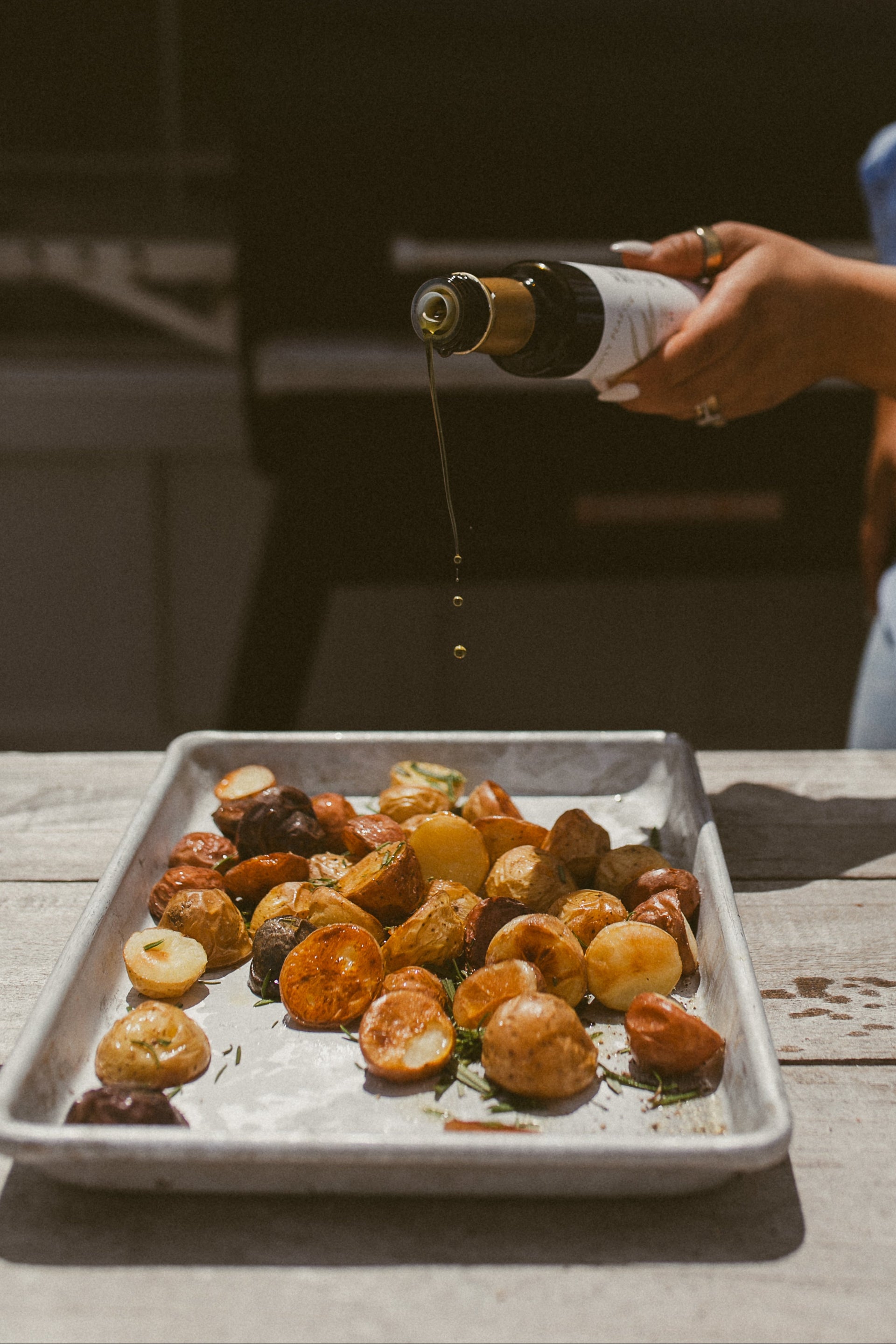 Person pouring olive oil over roasted vegetables on a baking tray outdoors.